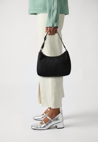 Borsa a mano in tessuto nero con silhouette curva e manico corto, tenuta in una mano. Accenti in tono argento su scarpe con tacco a blocco metallico e pantaloni chiari.