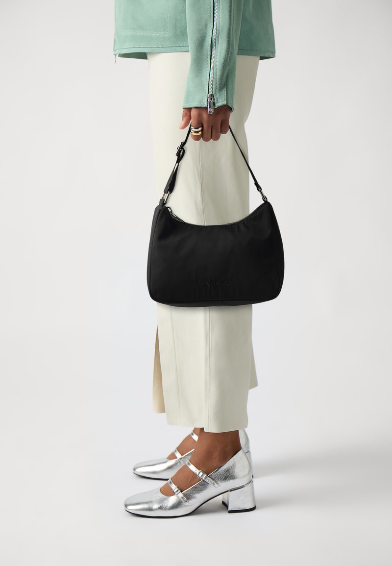 Borsa a mano in tessuto nero con silhouette curva e manico corto, tenuta in una mano. Accenti in tono argento su scarpe con tacco a blocco metallico e pantaloni chiari.