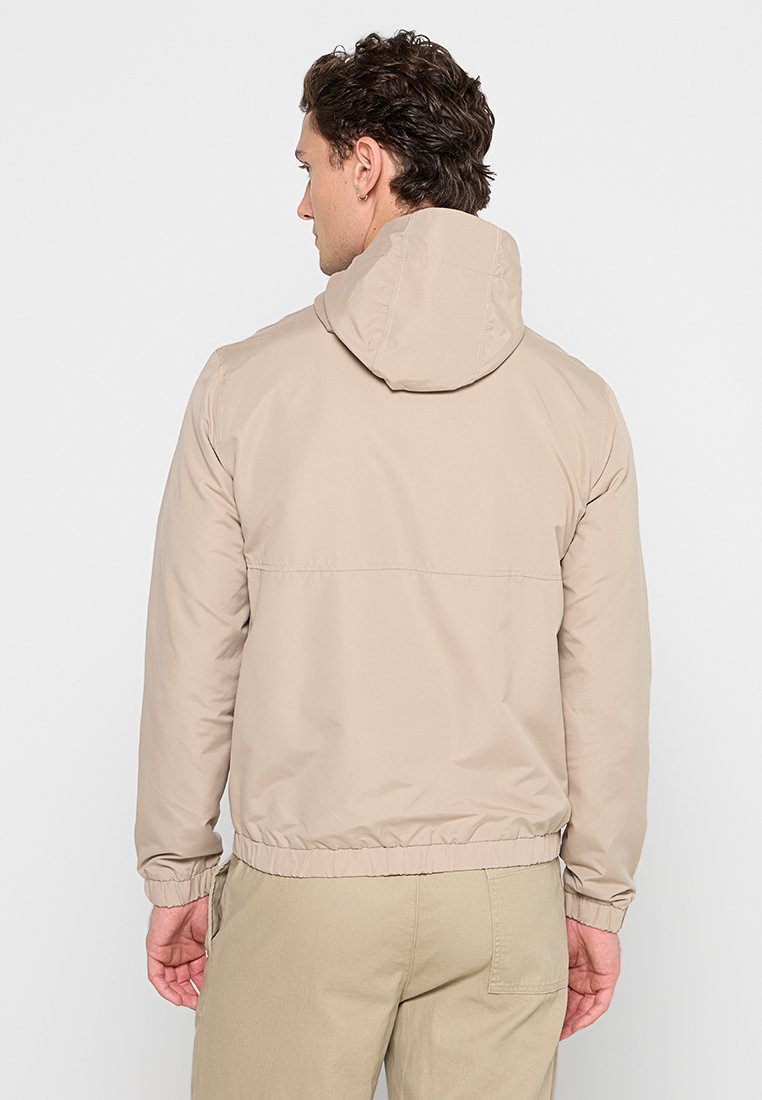 Chaqueta con capucha beige de textura suave, con puños y dobladillo elásticos. Diseño simple, sin patrones visibles ni detalles de hardware en la parte trasera.