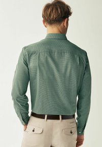 Grünes strukturiertes Hemd mit langen Ärmeln, mit Knopfmanschetten und Kentkragen, kombiniert mit hellen Hosen und einem braunen Gürtel.