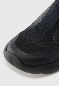 Zapatilla deportiva negra con parte superior de malla texturizada, banda elástica y suela de goma. Presenta una puntera lisa y brillante y un detalle sutil de la marca.