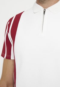 Homme portant un polo blanc avec une courte fermeture éclair argentée et des manches rouges comportant deux bandes blanches verticales.