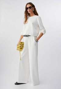 Sweat-shirt blanc avec détail brodé, associé à un pantalon blanc à jambes larges. Ceinture à pois noirs et blancs, pochette jaune et chaussures noires.