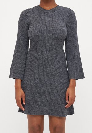 Kvinde iført en tætsiddende, ribstrikket grå kjole med trekvartlange klokkeærmer, stående mod en hvid baggrund.