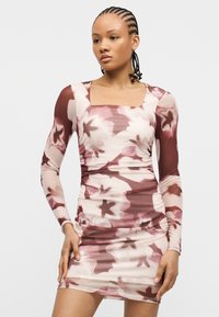 Γυναίκα με κοτσίδα που φοράει εφαρμοστό μακρυμάνικο φόρεμα με floral σχέδιο σε αποχρώσεις του ροζ και του βουργουνδίας, στέκεται μπροστά σε απλό φωτεινό φόντο.