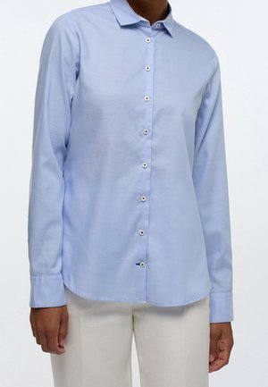 Osoba ubrana w jasnoniebieską koszulę na guziki z długim rękawem i kremowe spodnie, stojąca na tle jednolitej białej ściany.