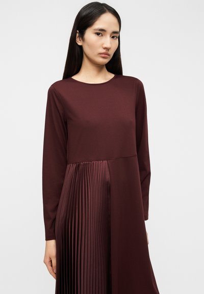 Το μπορντό φόρεμα με μακριά μανίκια διαθέτει λείο πάνω μέρος και πιετέ υφή στη φούστα, δημιουργώντας μια ρέουσα σιλουέτα με αντιθετικά τμήματα υφάσματος.