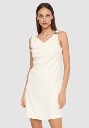 Donna che indossa un mini abito senza maniche color avorio con scollo drappeggiato e una fibbia circolare dorata su una spalla.