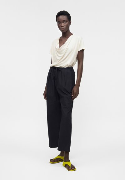 Γυναίκα στέκεται φορώντας ένα λευκό κοντομάνικο τοπ με draped ύφασμα, ψηλόμεσο μαύρο παντελόνι με φαρδιά πόδια και κίτρινα ανοιχτά σανδάλια σε λευκό φόντο.