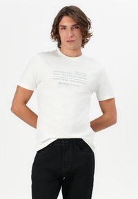 Camiseta de algodón blanca con mangas cortas que presenta un texto impreso en gris y azul. Combinada con unos vaqueros de denim negros. Diseño simple y casual.