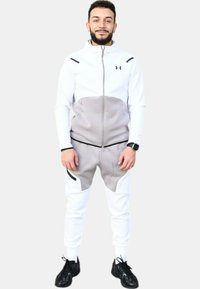 Chándal deportivo blanco y gris con chaqueta con cremallera que presenta acentos en los hombros, combinado con pantalones ajustados; llevado con zapatillas negras.