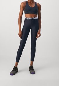 Granatowy stanik sportowy i legginsy z wysokim stanem, obie części dobrze dopasowane. Legginsy mają logo "UNDER ARMOUR" na pasie i małe logo na nogawce.