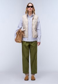 Beige padded vest over a blue and white striped shirt, olive green pants, and brown slip-on shoes. Holding a tan bag. Sunglasses worn.