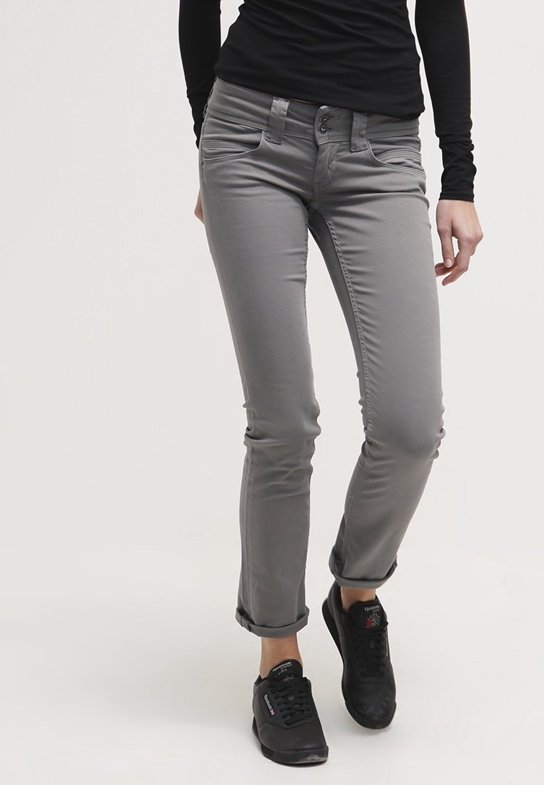 Personne portant un pantalon gris ajusté avec les revers roulés et des baskets noires Reebok, debout devant un fond uni.