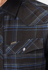 Camisa de cuadros en negro y azul con una tela texturizada. Cuenta con un bolsillo en el pecho con botón y una etiqueta con el logo. Cuello clásico y detalles de costura en contraste.