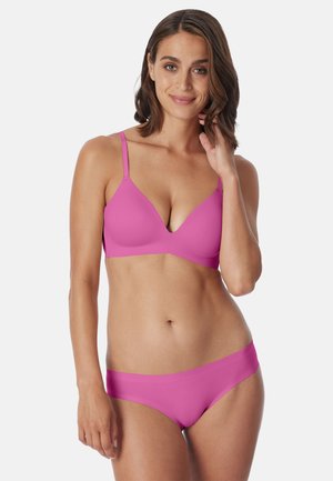 Woman smiling, wearing matching pink bra and underwear set, with short wavy hair and light background.