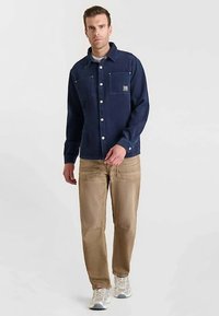 Camisa azul marino abotonada con dos bolsillos, elaborada con tela suave. Combinada con pantalones marrón claro. Zapatillas blancas y grises completan el conjunto.