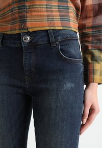 Mörkblå denimjeans med klassisk passform, framfickor och silverfärgad knäppning. Tyget har en subtil textur och lätt blekning.