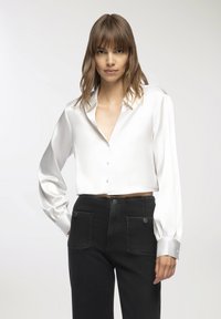 Blusa bianca in satin, tagliata, con scollo a V, maniche lunghe a sbuffo e polsini con bottoni. Indossata con pantaloni in denim neri a vita alta con tasche.