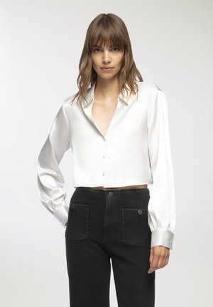 Blusa bianca in satin, tagliata, con scollo a V, maniche lunghe a sbuffo e polsini con bottoni. Indossata con pantaloni in denim neri a vita alta con tasche.