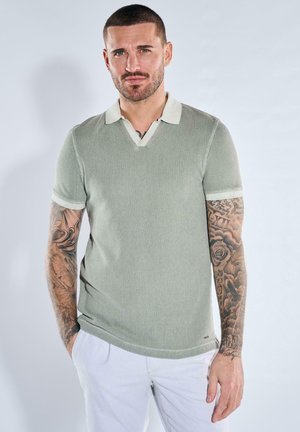 Mann mit kurzen Haaren und Tätowierungen auf beiden Armen, trägt ein hellgraues, strukturiertes Poloshirt und weiße Hosen, steht mit den Händen in den Taschen.