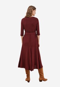 Bordeauxrotes Langarmkleid mit taillierter Oberseite, ausgestelltem Rock und einem gebundenen Gürtel in der Taille. Getragen mit knielangen, braunen Stiefeln.