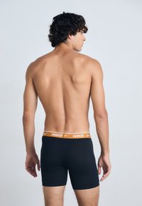 Črne Nike bokserice z oranžnim pasom, ki se prilegajo in imajo gladko teksturo. Pogledujejo s hrbta, model ima kodraste lase.