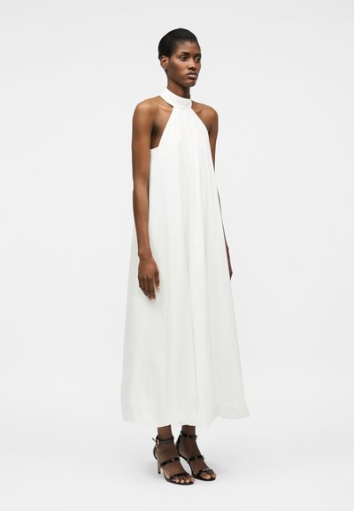 Femme portant une robe longue blanche sans manches à col licou, avec des sandales noires à talons hauts avec lanières, debout devant un fond uni.