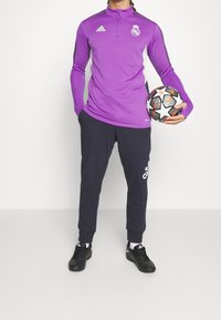 Lila träningsöverdel med långa ärmar och zip-up krage med vita accenter, matchad med mörkblå joggingbyxor och svarta fotbollsskor, håller en fotboll.