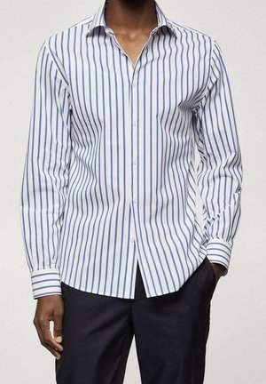 Mężczyzna w białej, długorękawowej koszuli z pionowymi niebieskimi paskami i ciemnych spodniach, jedna ręka w kieszeni, zwrócony przodem.