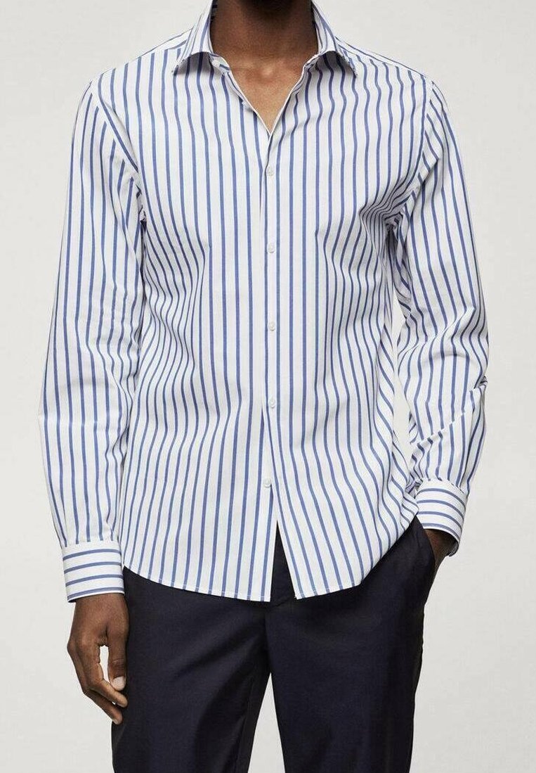 Homme portant une chemise blanche à manches longues avec des rayures verticales bleues et un pantalon foncé, une main dans la poche, faisant face à l'avant.