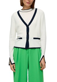Witte cardigan met een marineblauwe afwerking, V-hals ontwerp en knopen aan de voorkant, gecombineerd met groene wijde broekspijpen. De stof lijkt glad.
