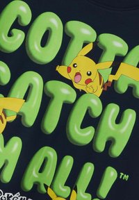 Juodos spalvos marškinėliai su didelėmis, blizgiomis žalios spalvos raidėmis, skaitančiomis "PRIVALAU SURINKTI JUOS VISUS!", ir dviem Pikachu iliustracijomis geltonais, juodais ir raudonais akcentais.