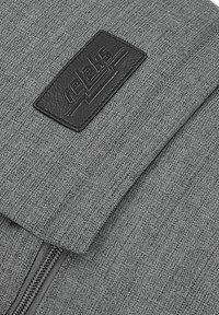 Sac à dos en textile gris avec un patch logo en cuir noir bien en vue, doté d'une fermeture éclair et d'une surface en tissu texturé.
