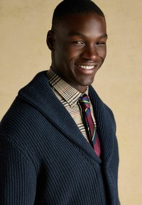 Marineblauer, voluminöser Strickcardigan mit Revers, über einem karierten Hemd und einer gestreiften Krawatte getragen. Glatte Haut und kurzes, dunkles Haar.