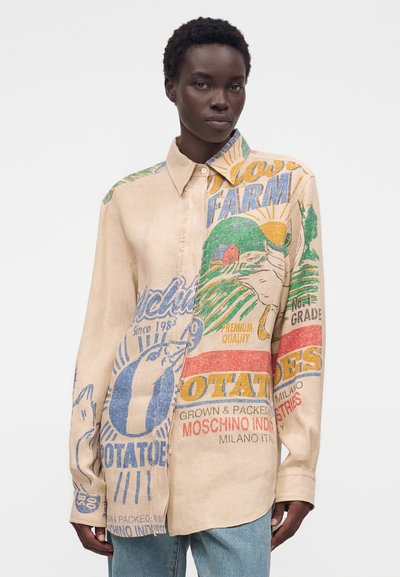 Γυναίκα μοντέλο φοράει μπλούζα μπεζ με πολύχρωμο vintage σχέδιο από σάκο πατάτας και κείμενο, σε συνδυασμό με ανοιχτό μπλε τζιν, σε λευκό φόντο.