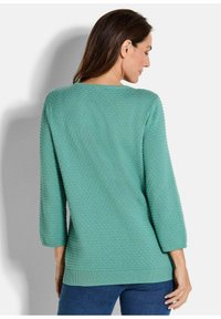 Frau in einem strukturierten türkisfarbenen Strickpullover mit dreiviertelÄrmeln, von hinten vor einem weißen Hintergrund dargestellt.