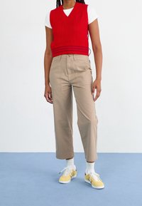 Rode gebreide mouwloze trui over een wit T-shirt; beige wijde broek en gele sneakers. Witte sokken met bruine accenten.