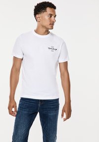 Vit bomullströja med korta ärmar, med en svart tryckt logotyp på vänster bröstficka; bärs med mörkblå jeans.