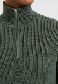 Maglione a coste di un verde scuro, con collo alto e mezza zip. Presenta un tessuto texturizzato con righe orizzontali.