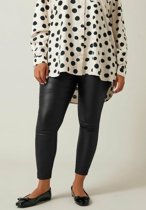 Witte blouse met grote zwarte stippen, gecombineerd met glanzende zwarte legging. Zwarte flats met een strikaccent maken de outfit compleet.