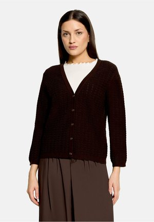 Woman wearing a dark brown buttoned cardigan over a white top with a scalloped neckline and matching brown pleated skirt.