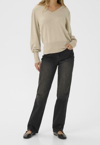 Pull beige à col en V avec manches longues légèrement bouffantes, associé à un jean noir à jambes larges et des chaussures plates noires. Look simple et décontracté.