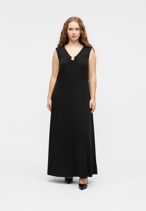 Γυναίκα με μακριά σγουρά μαλλιά, που φοράει μαύρο μακρύ αμάνικο φόρεμα με άνοιγμα στο ντεκολτέ και μαύρες μυτερές γόβες, όρθια.