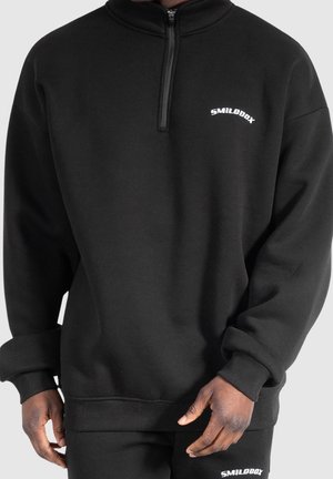 Persoon draagt een zwarte trui met een kwart ritssluiting en bijpassende broek met witte "SMILODOX" logo's op de borst en dij, handen zichtbaar aan de zijkanten.