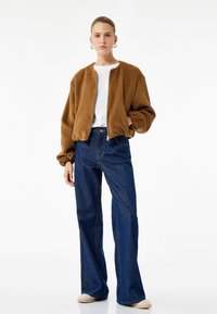 Veste bomber en velours côtelé brun avec zip, associée à un jean évasé bleu taille haute et un t-shirt blanc ; modèle portant des chaussures dans des tons neutres.