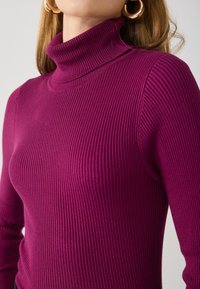 Tettsiddende ribbestrikket turtleneck-genser i dyp magenta med teksturert stoff. Har høy hals og lange ermer.