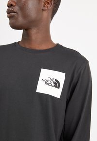 Schwarzes langärmliges T-Shirt aus Baumwolle, mit einem weißen rechteckigen Logo-Patch, auf dem der Text "THE NORTH FACE" in Schwarz zu sehen ist.