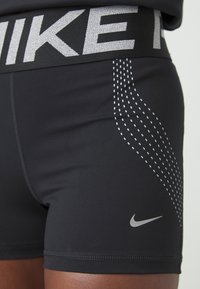 Gros plan sur un short noir Nike avec des lignes blanches ponctuées et le logo réfléchissant Nike swoosh sur la cuisse, ainsi qu'une ceinture Nike large.