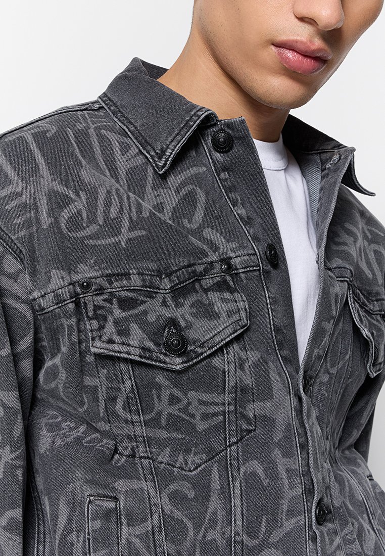 Chaqueta de mezclilla gris con patrones llamativos en estilo graffiti negro, bolsillos con botón y un cuello clásico. El tejido texturizado añade profundidad.